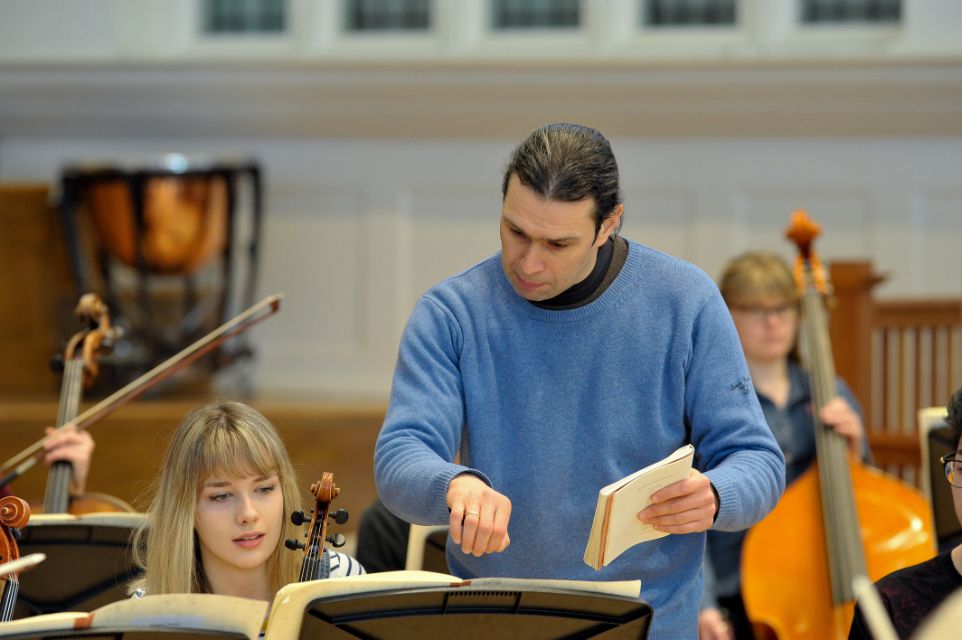 Man, wearing a blue shirt, talking to a student, sitting in a orchestra rehearsal.